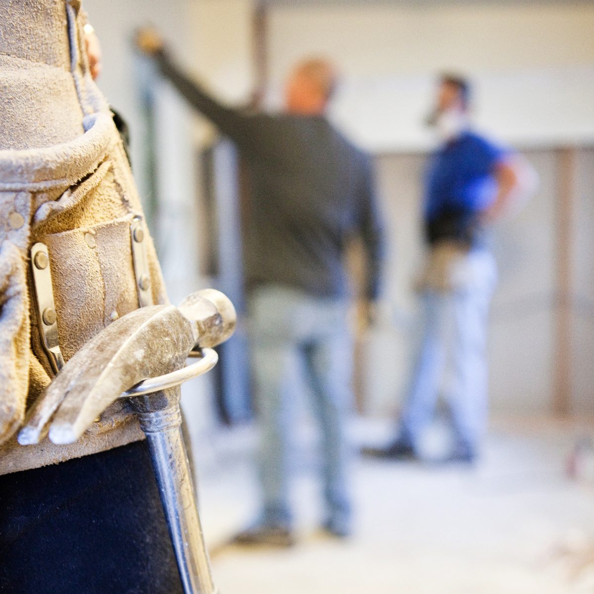 A contractor tool belt with a hammer, with two tradesmen working in the background